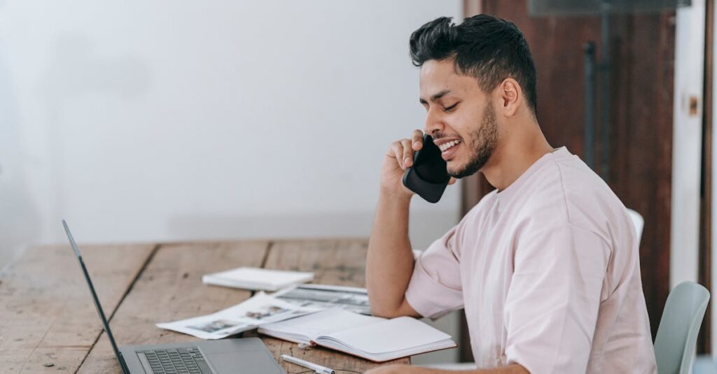Young Hispanic man engaged in a phone call while working remotely on a laptop at home.